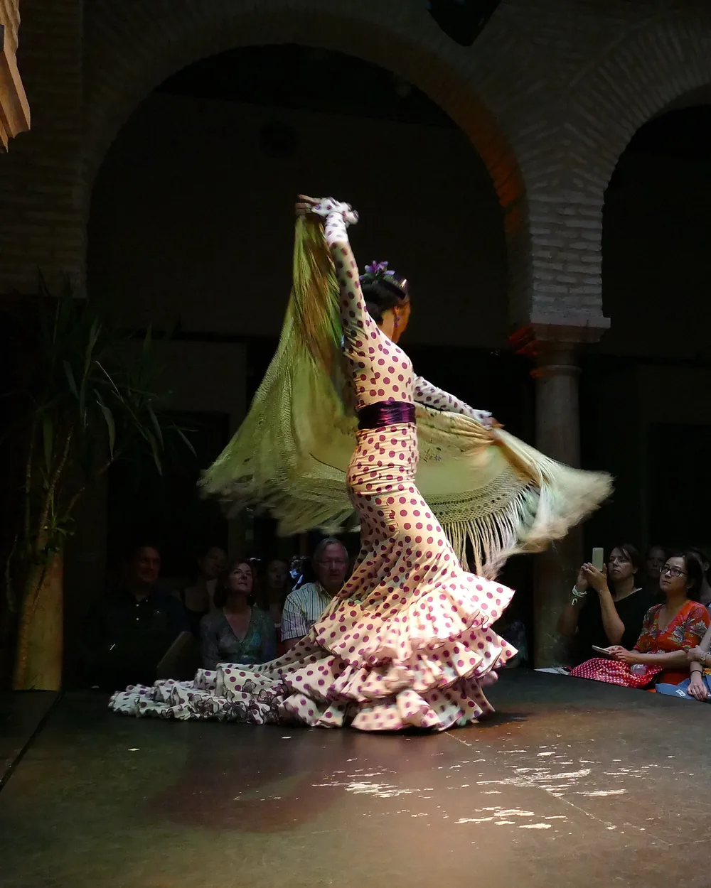 A flamenco dancer in a polka-dot dress turns with a long fringed shawl on the museum's low stage, with spectators seated on the right and brick arches behind