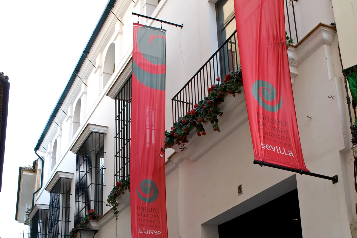 The white street front of the Museo del Baile Flamenco with red banners, a balcony of red flowers and a dark open doorway