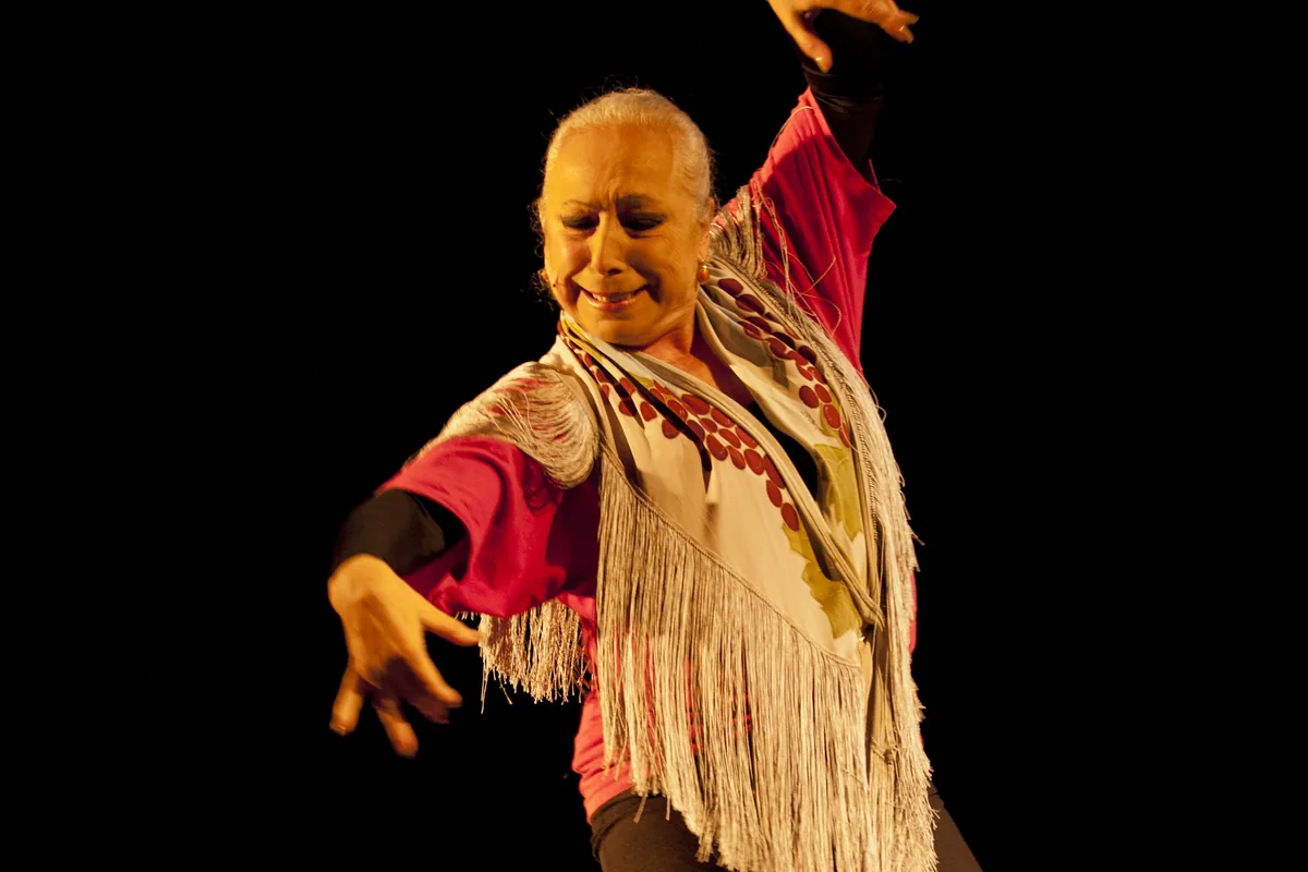 Cristina Hoyos on a dark stage in a red top, with a long fringed shawl over her shoulders and one arm raised, smiling as she dances